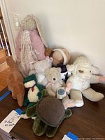 Group photo of nine assorted stuffed animals including Floppy Tomkins bunny, JB Bean bear, lambs, turtle, and others arranged together on a brown cloth.