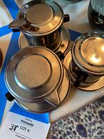 Top-down view of two stainless steel Vietnamese phin coffee makers placed on their saucers, showing lids and black handles.