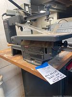 Scroll saw mounted on left side of wooden work table with blue tag showing lot and item numbers visible.