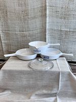 Three Corning Ware white saucepans with handles, two with blue cornflower patterns. One saucepan has a spout. One clear glass lid in front.