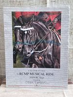 Full view of the 2009 RCMP Musical Ride BC Tour print showing two black horses with riders, text and signature of Joan Larson. Visible water damage partially veiled by thin blue line.
