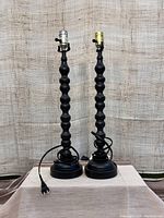Pair of two modern black metal table lamps with cords, placed on white surface in front of beige background.
