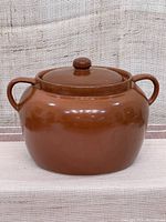 Front view of vintage brown ceramic bean pot with lid and two side handles on a fabric background.