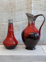 Two ceramic pottery pieces, a large pitcher and a smaller narrow-necked vase, shown side by side on white fabric with a natural textured background. Both have red to black drip glaze finish.