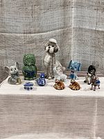 Front view of all 11 ceramic and glass animal figurines arranged in a line on a textured cloth surface. Includes owl coin bank, dog, elephants, mice, and birds.