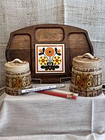 Wooden 1970s cutting board with carved compartments and decorative floral tile, alongside ceramic canisters and candles in front