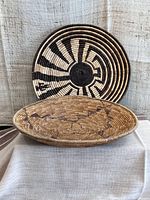 Pair of large shallow woven baskets displayed upright and stacked, showing contrasting light and dark woven patterns.