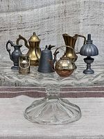 Full set of 7 vintage metal miniatures displayed on a glass cake stand, showing pitchers, kettles, lamp, and milk jug