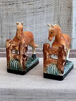 Pair of ceramic horse bookends shown at angle with brown horse standing by rustic fence on green grass base.