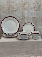 Set of six white ceramic dishes with burgundy rim accents, including cups, saucer, side plate, and dinner plates, arranged on a surface against a fabric background.