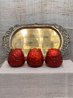 Three red textured glass table candle holders in front of a heavy brass serving tray with ornate rim, showing frontal view.