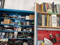 Overview photo showing blue shelving unit filled with assorted automotive parts, wiring spools, various boxed items, and a separate wooden shelf holding automotive manuals and catalogs.