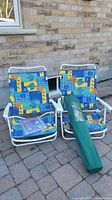 Two folding beach chairs with colorful fabric and white frames positioned outside on a patio with next to a brick wall. Green carrying bag chair placed on one chair. Some chair covers visible in background.