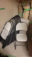 Stack of 13 white resin folding chairs with black metal frames leaning against wall. Rolled carpets in background.