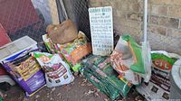 Wide angle photo showing collection of lawn and garden soil, manure, Turf Builder fertilizer bags, garden sign leaning against wall, and a white garden sprayer on left side.