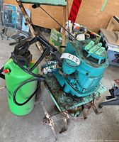 Photo shows the teal Yuba rototiller with attached handles and black cables next to the green Home Gardener insecticide sprayer with pump handle and hose.