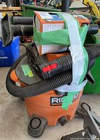 Ridgid shop vac unit with black hoses and nozzle attachments taped on top and around the unit, showing the front and side of the orange body with logo visible.