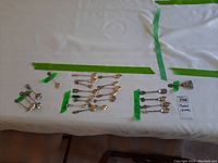 Wide view of multiple souvenir spoons arranged in groups, each group separated with green tape, showing variety of spoons.