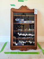Front view of the wooden display case showing multiple souvenir spoons inside against black felt backing.