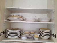 Three-shelf cabinet view showing stacks of plates, bowls, mugs, creamers and spoons