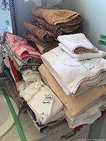 Stack of assorted towels including beige and brown bath towels, white hand towels with leaf embroidery, and various colored towels stacked neatly.