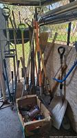 A group of yard and garden tools including rakes, shovels, broom, and a box containing small garden tools on the floor