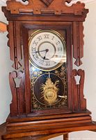 Front view of the antique gingerbread clock showing carved wood frame, Roman numeral dial, glass panel with gold decoration, and pendulum.