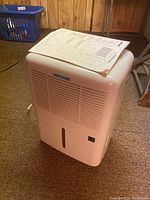 Front view of white Arcticaire portable dehumidifier with manual on top on brown carpet basement floor.