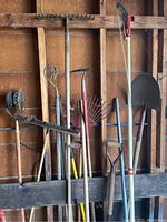 Full view of assorted garden tools including rakes, a shovel, a tiller, a branch trimmer, and brooms positioned side by side against a wooden wall.