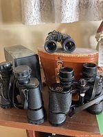 Three sets of binoculars displayed with a brown leather carrying case on a shelf.