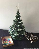 Full view of the ceramic Christmas tree with white snow accents, clear star topper, electrical cord, and a box of multi-colored plastic lights on a black background.