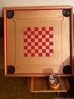 Full view of the wooden game board, showing the checkerboard pattern with red squares and four black mesh pockets in the corners. Tin container with checkers set on bottom right side.