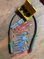 Photo showing flexible drill extension, yellow hole saw storage box, and various drill bits in packaging laid out on wooden floor