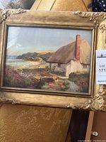 Full view of framed coastal cottage landscape painting showing gold ornate frame and coastal scenery with cottage and garden.