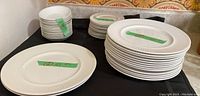 Wide shot showing stacks of white ironstone plates and bowls on a black cloth surface, labeled with green tape indicating size.