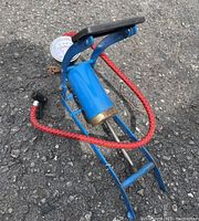 Blue foot pump with a mechanical pressure gauge attached and a red braided hose on asphalt surface.