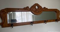 Full view of the rectangular mirror with wooden carved frame and four black metal hooks beneath it.