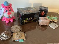 Full lot overview showing pink bear money bank, black metal safe bank, ornate jewelry box, painted floral jewelry box, three belt buckles, silver compact, and lipstick holder.