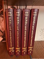 Four maroon hardcover books with gold embossed spines authored by Yves Naud titled 'UFOs and Extraterrestrials', volumes 1 to 4, arranged vertically on a wooden shelf.