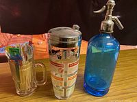 Photo showing clear glass beer stein filled with multi-colored drink sticks and packets, next to a blue glass seltzer bottle with metal spout, and a tall cocktail shaker with printed recipes.