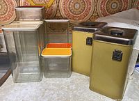View of 2 yellow metal canisters with stainless steel lids and 7 assorted plastic canisters with colored lids on kitchen counter, vintage style background.