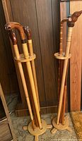 Photo showing six wooden walking canes with a variety of carved handles including animal head designs, held upright in two round wooden stands made of light wood.