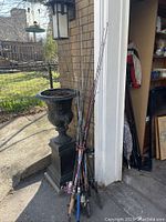 Photo showing a cluster of fishing rods leaning against a house corner next to a black ornamental planter. Rods vary in handle colors including pink, blue, orange, and black.