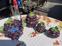 Full view of antique grape ceramic set items on a floral tablecloth under sunlight showing the cookie jar, teapot, sugar bowl, and marmalade jar shaped and decorated like grape clusters with green leaves.