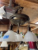 Side angle showing the fixture hanging, three white milk glass shades with scalloped edges, solid brass base and chain, garage background.