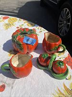 Photo of antique tomato-shaped ceramic set including sugar bowl, creamers, and salt and pepper shakers on a floral tablecloth outdoors.