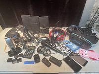 Photo showing a large assortment of untested electronics including two Apple iPhones, multiple headphones, speakers, shavers, remote controls, and accessories on a table.