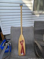 Full front view of the wooden canoe paddle against a chair and siding background, showing length and handle shape.