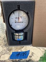 DrumDial dial gauge in foam packaging inside open box, showing white face and black numerals.