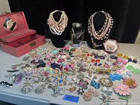 Photo showing a large selection of costume jewelry laid out on a table including colorful stud earrings, multi-strand bead necklaces on busts, brooches, and a pink wooden jewelry box.